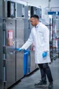 Technician in a white lab coat handling an aging test chamber in a laboratory for medical device packaging validation.