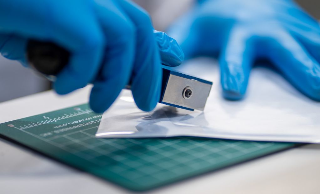 A laboratory technician wearing blue gloves conducting seal integrity testing on medical packaging using a precision cutting tool.