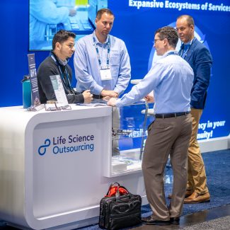 Two men shaking hands at a "Life Science Outsourcing" booth, with two onlookers, at a business expo.