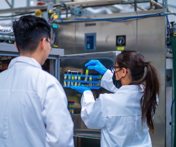 Two laboratory technicians in white lab coats conducting bioburden testing, handling test tubes with liquid samples near a sterilization chamber in a controlled lab setting.