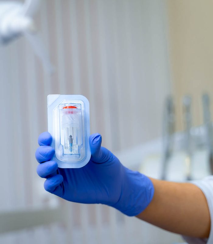 Dental,Implant,In,Dentist`s,Hands.,Modern,Dentistry,Concept.,Blurred,Background. Gloved hand holding a sterile medical device in clear protective packaging, emphasizing safety and quality in healthcare.
