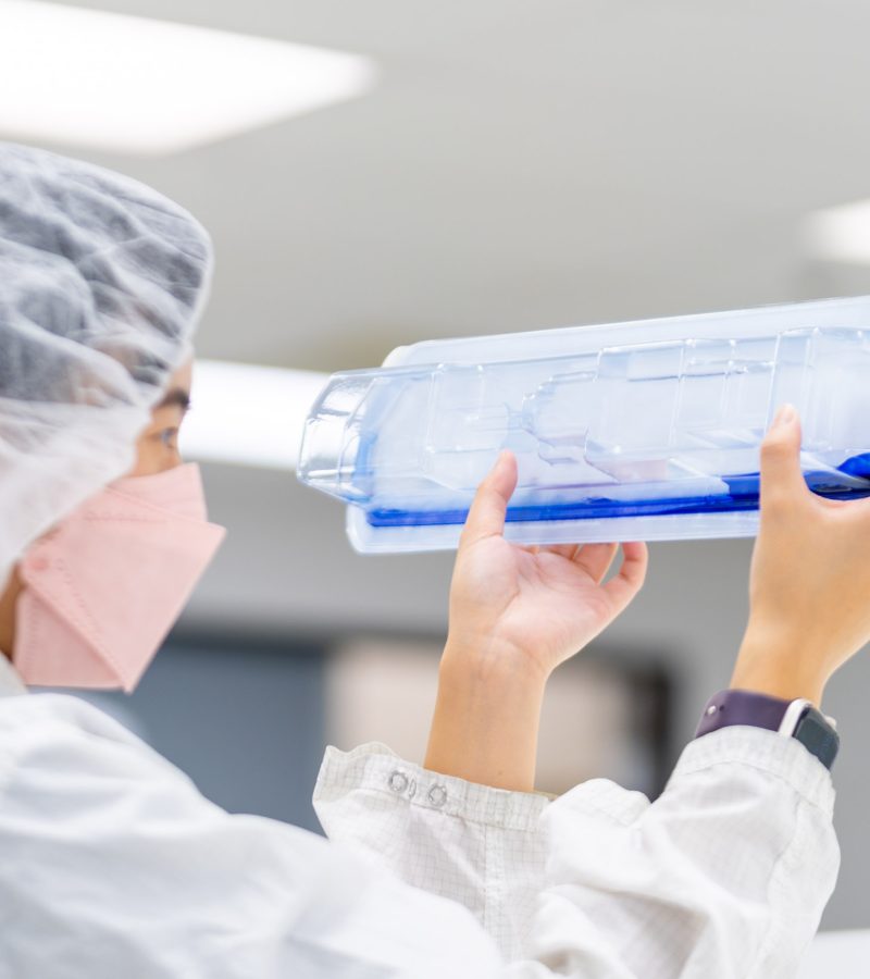 Technician wearing protective gear closely examining medical device packaging for leaks during a dye test in a laboratory setting.