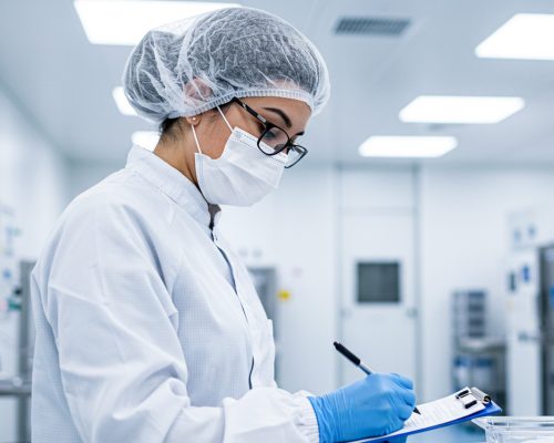 Quality Control Inspection in a Cleanroom Environment A laboratory technician in cleanroom attire conducts a quality control inspection with a clipboard and pen in a sterile facility.