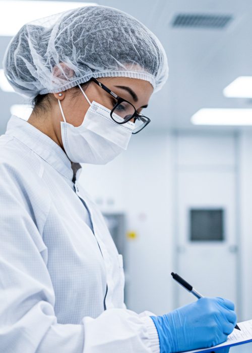 A laboratory technician in cleanroom attire conducts a quality control inspection with a clipboard and pen in a sterile facility.