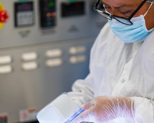 Technician wearing gloves and a mask inspecting medical device packaging on a light table in a quality control lab.