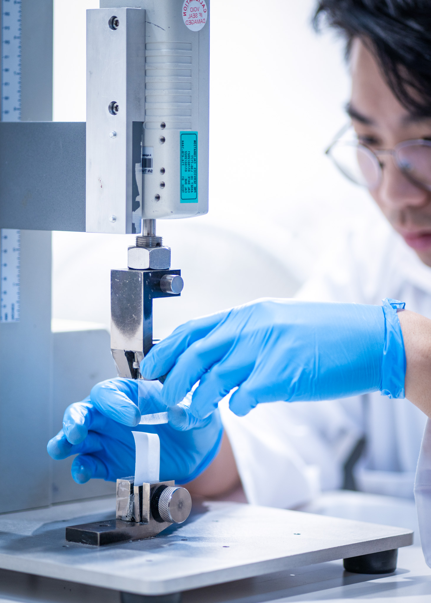 A laboratory technician in blue gloves conducting a peel strength test on medical packaging using a tensile testing machine in a controlled lab environment.