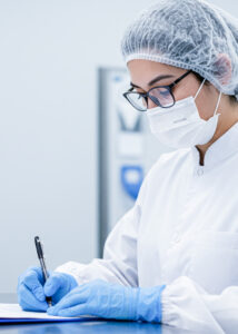 Technician in a cleanroom writing compliance documentation while wearing protective gear, including gloves, a mask, and a hairnet.
