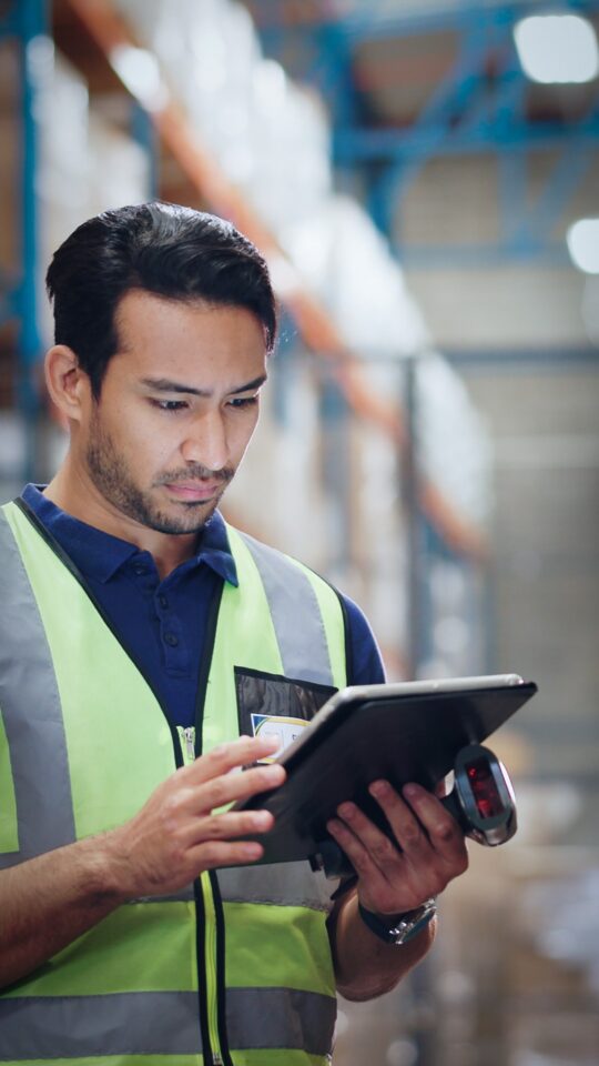 Warehouse worker in a safety vest using a tablet and barcode scanner for inventory tracking